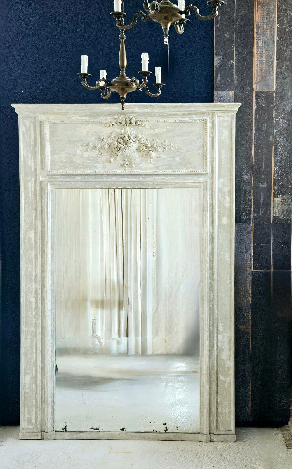 A large, distressed white fireplace mirror with an ornate carved frame, set against a dark navy blue wall and a dark wood-paneled wall. A brass candelabra hangs above the mirror.